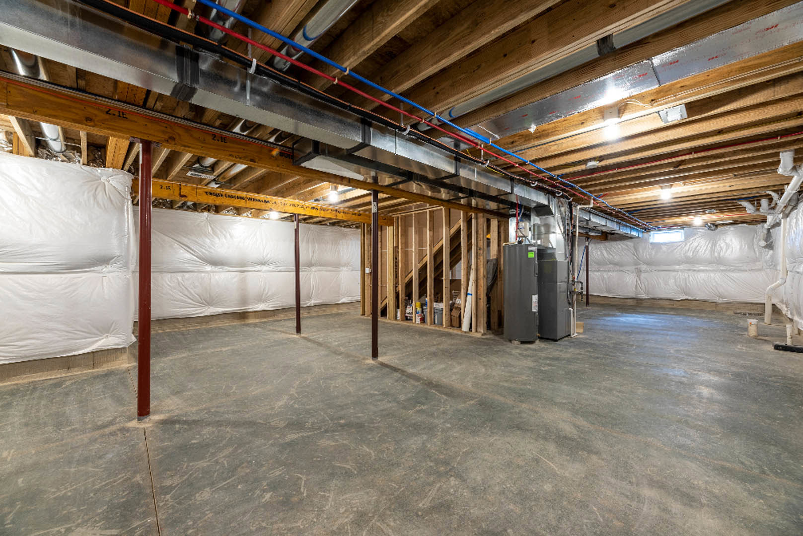 Spacious basement with exposed steel pipes and beams on a high ceiling, concrete floor, wood framing, white insulation sheets, black electrical box, and door detail