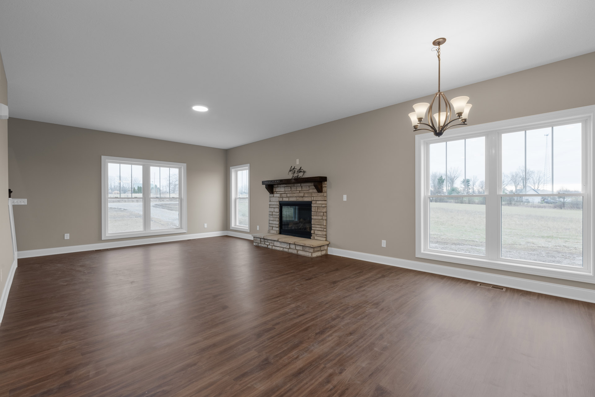 Large living room with hardwood floors, white plaster walls, central fireplace featuring a wood mantel shelf, and windows overlooking a dirt road and neighboring house