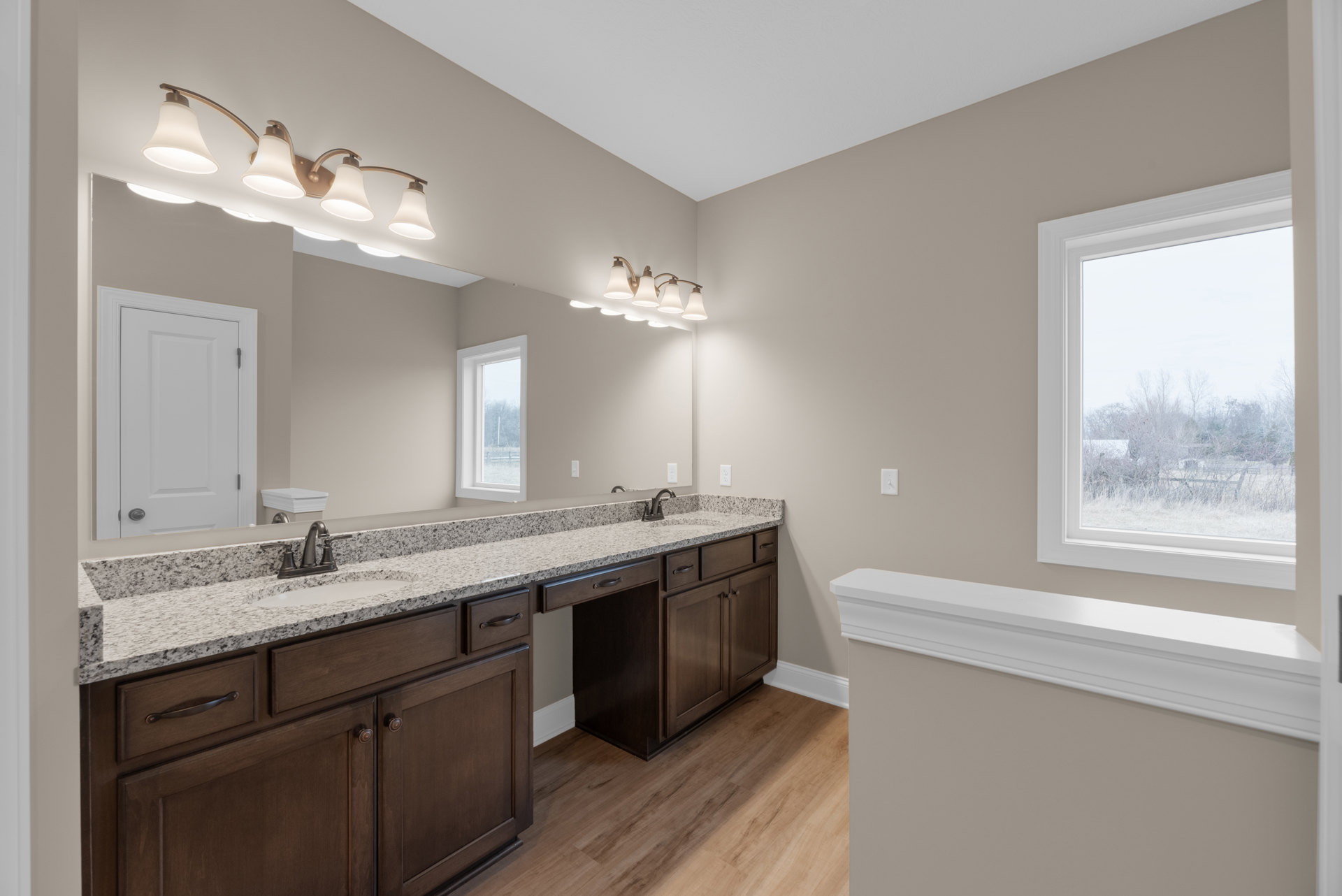 Bathroom with marble countertop, double sinks, large framed mirror, white cabinetry, black hardware, window overlooking field and trees, white light fixture with shade.