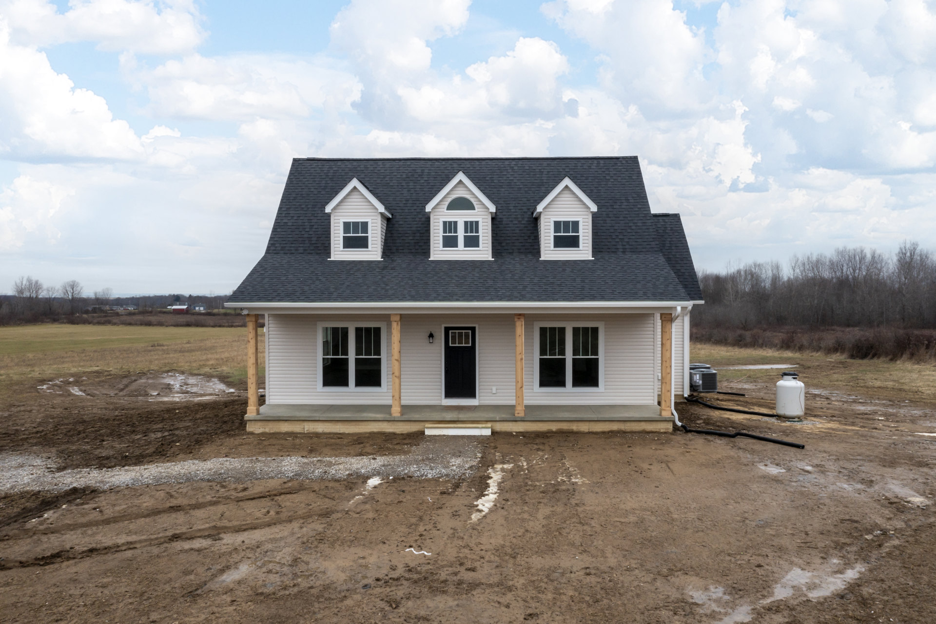 Two-story house under construction with white siding, black door, white-framed windows, dormer roof, dirt yard, and cloudy sky