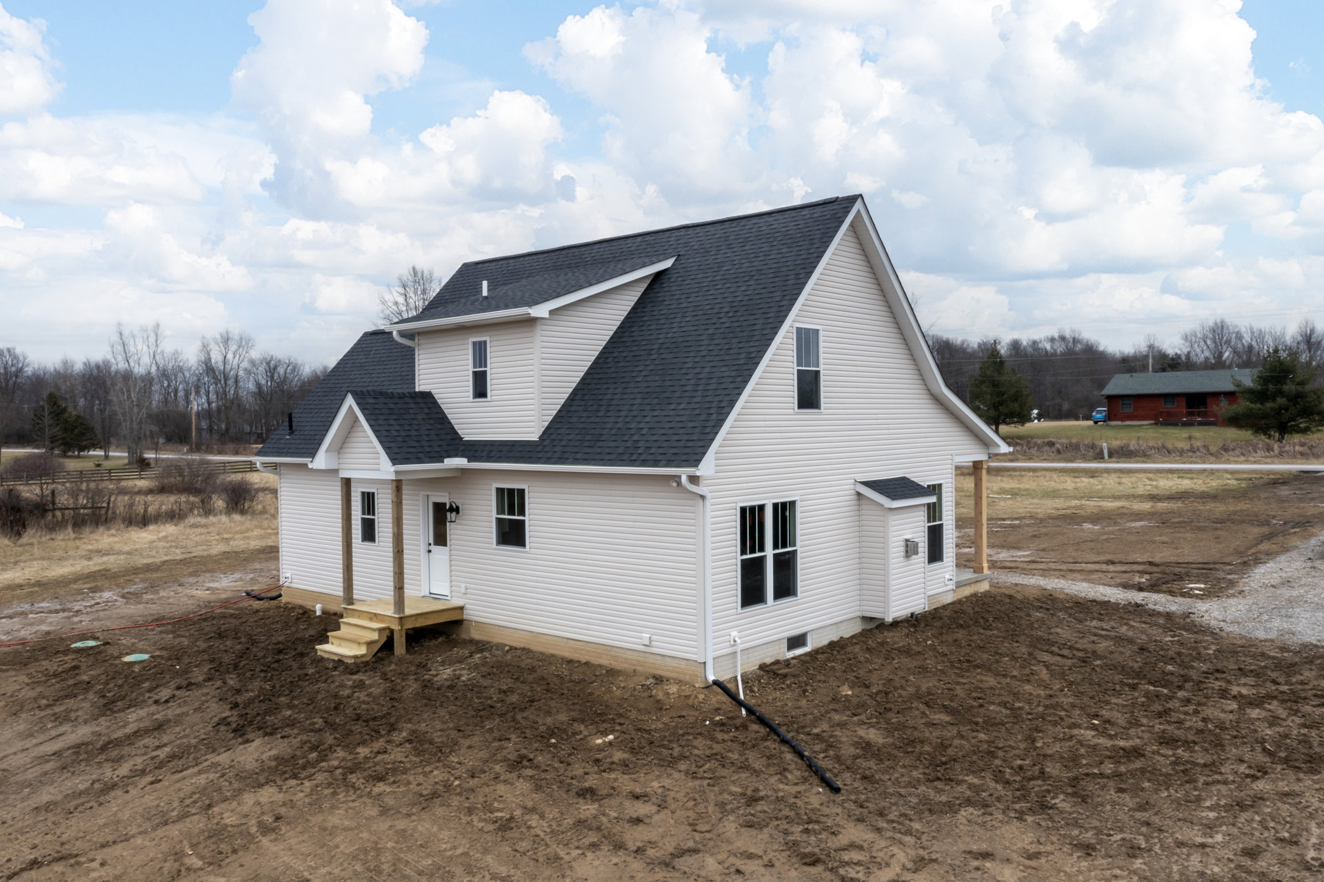 Two-story house under construction with exposed framing, covered porch, wooden deck, dirt yard, scattered construction materials, black pipe on ground, mature trees in background