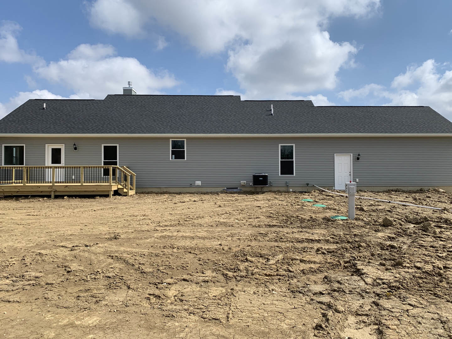 Modern house with light siding, white-framed window, wooden deck featuring metal railing, dirt yard bordered by a metal wall, blue sky with scattered clouds