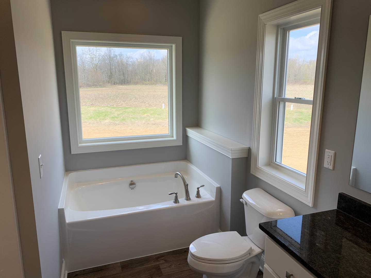Bathroom with white tile walls, freestanding bathtub beneath a window overlooking a grassy field, black granite countertop with sink and chrome faucet, toilet adjacent to tub.