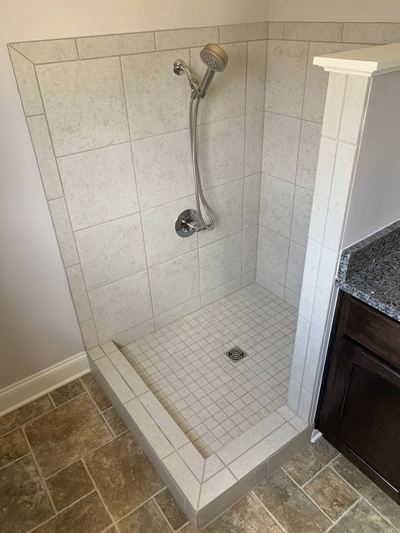 Walk-in shower with chrome shower head mounted on white tile wall, linear drain on tile floor, partial view of glass door, granite countertop adjacent to shower.