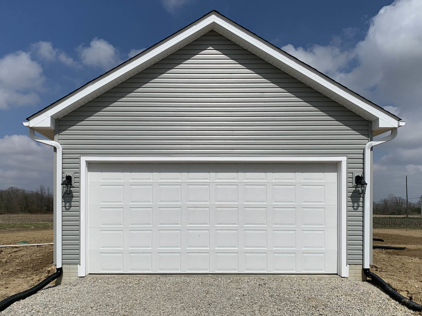 White paneled garage door beneath gray shingle roof, gravel driveway in foreground, light siding, trees in background