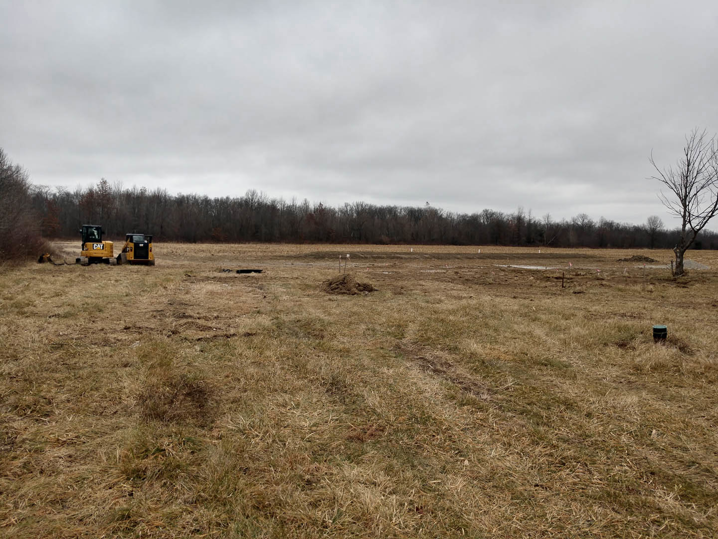 Yellow tractor parked on grassy field beside pile of dirt, leafless tree in background, cloudy sky overhead, rural landscape with scattered trees.