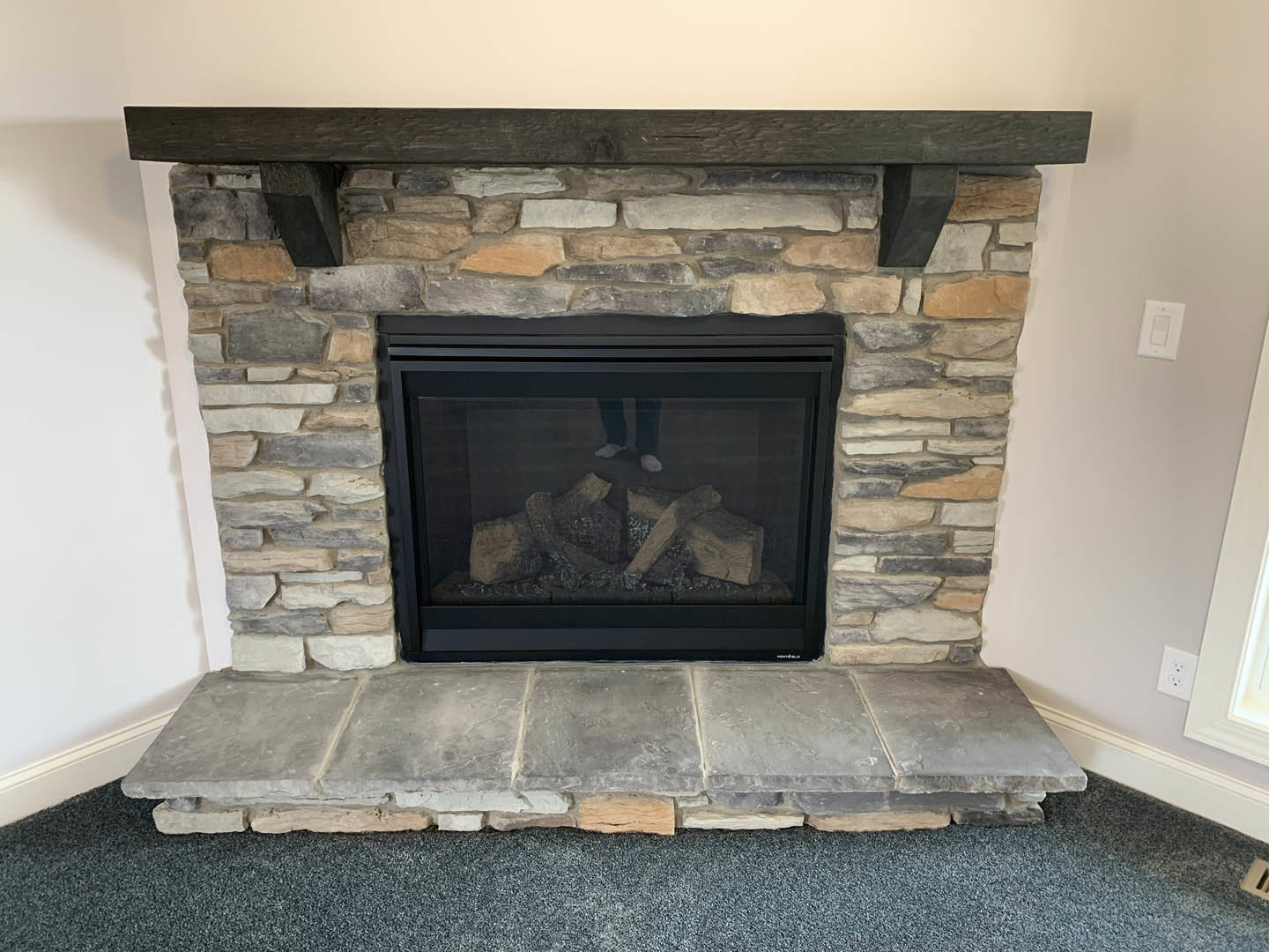 Stone fireplace with glass door, wood mantle shelf, white outlet and light switch on adjacent wall, hardwood floor