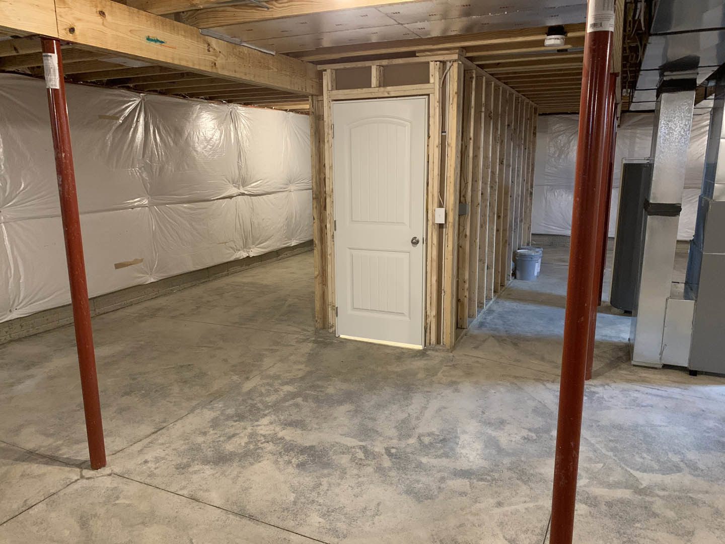 White door with silver knob set in a wall covered in plastic sheeting, paint bucket on floor, exposed metal ceiling panel, and close-up of support pole in unfinished basement