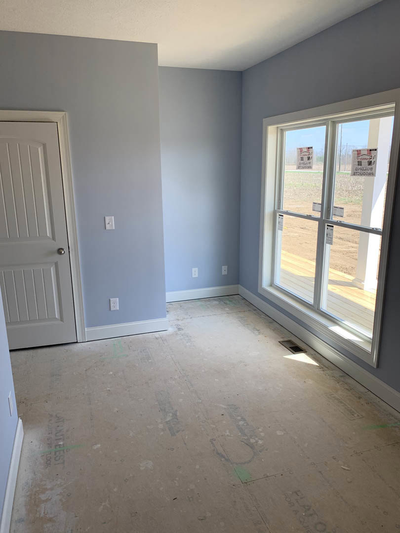 White plaster walls, wood flooring, single-pane window with building company sign, modern door with black hardware, natural light casting shadows across interior room