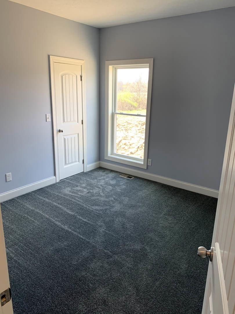 Neutral-toned carpeted room with white plaster walls, large window offering outdoor view, and white paneled door with silver knob