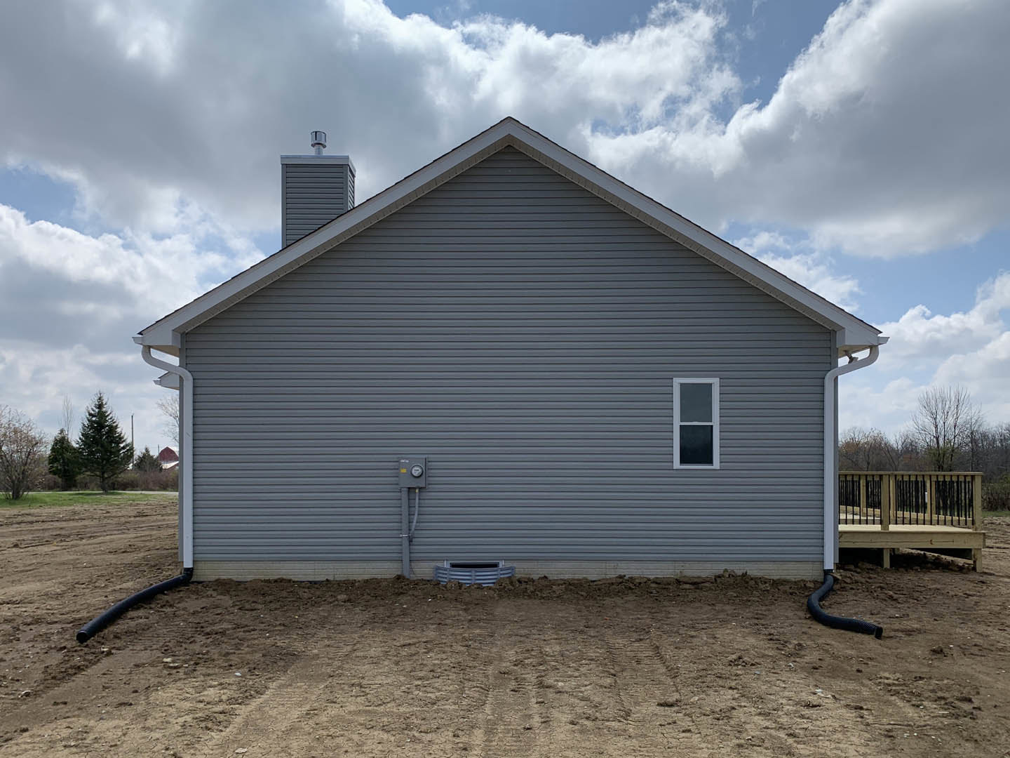 Two-story house with light siding, black-railed wooden deck, white-framed windows, fenced yard, and chimney, set against cloudy sky and dirt ground.