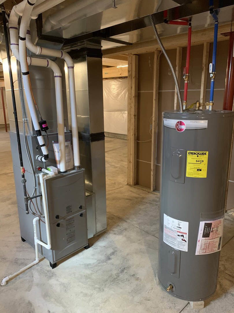 Large grey water heater tank with yellow and white label, connected to pipes and tubes, set against an indoor wall with visible floor and fire extinguisher nearby.