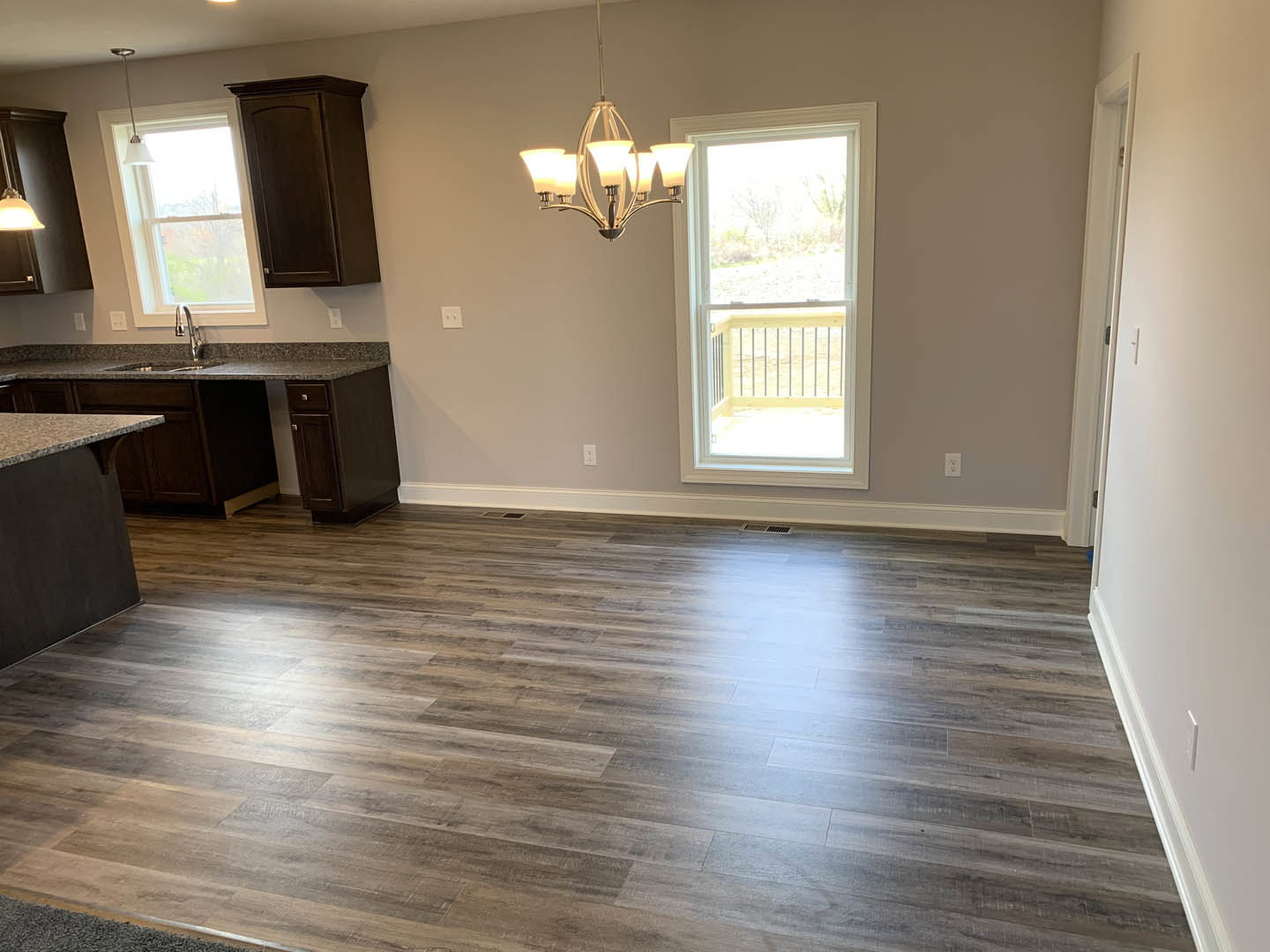 Open kitchen and dining area with hardwood flooring, brown cabinetry, ceiling chandelier, and large window with railing.
