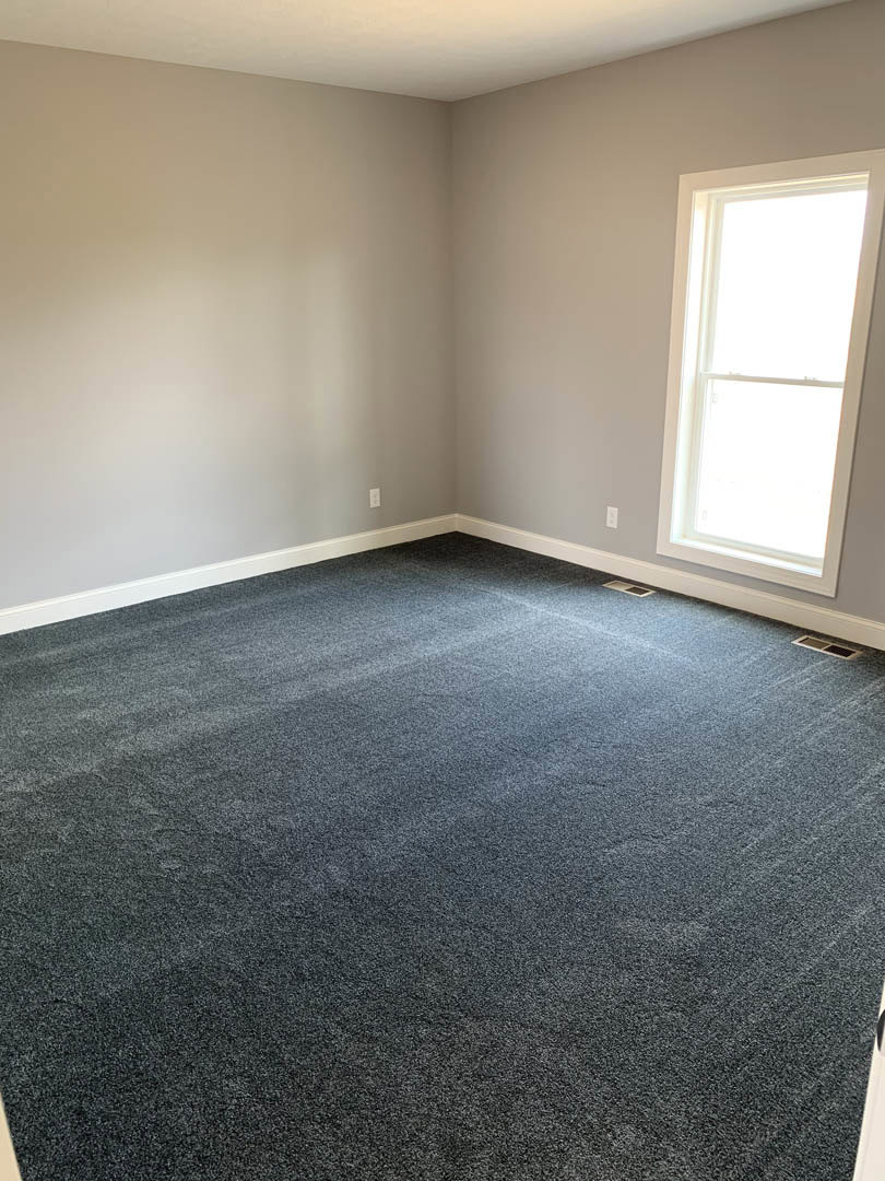 Carpeted room with white walls, large window with white frame, blue sky and clouds visible outside
