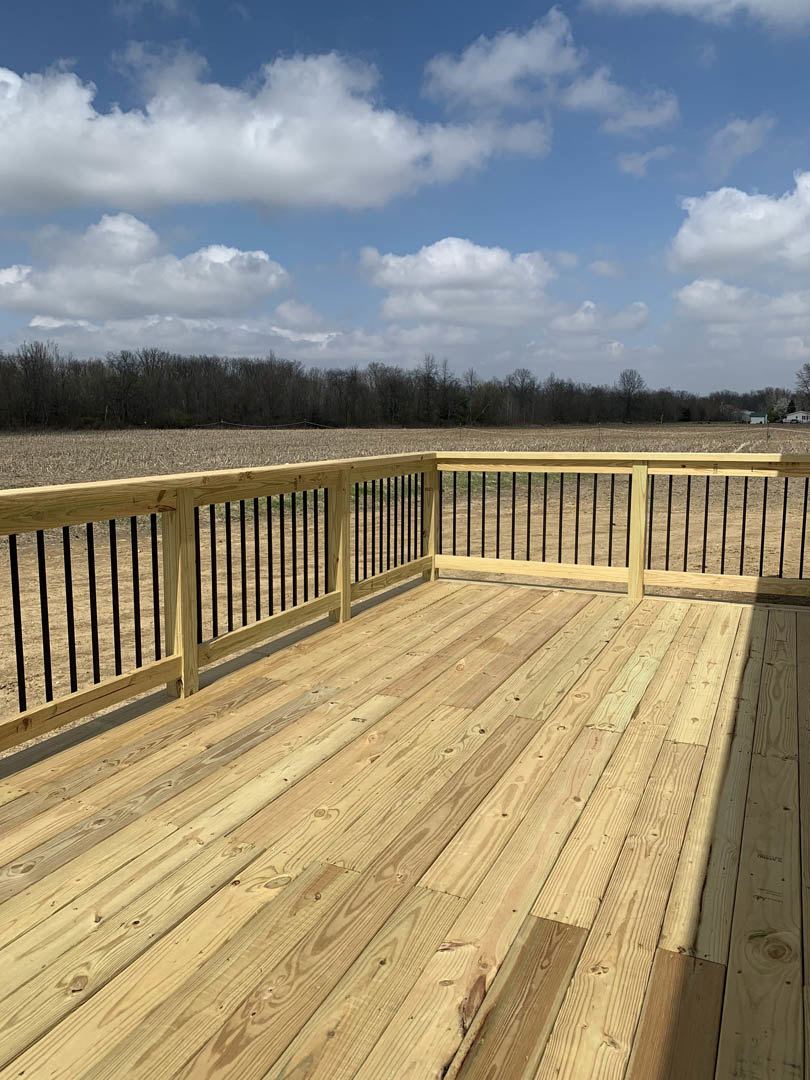 Wooden deck with black metal railings overlooking a field of dense trees under a cloudy sky