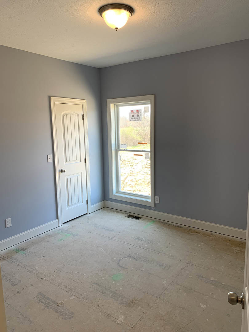 White paneled door and rectangular window in a plaster-walled room, ceiling-mounted light fixture, unfinished flooring with visible hole, natural light entering from outside