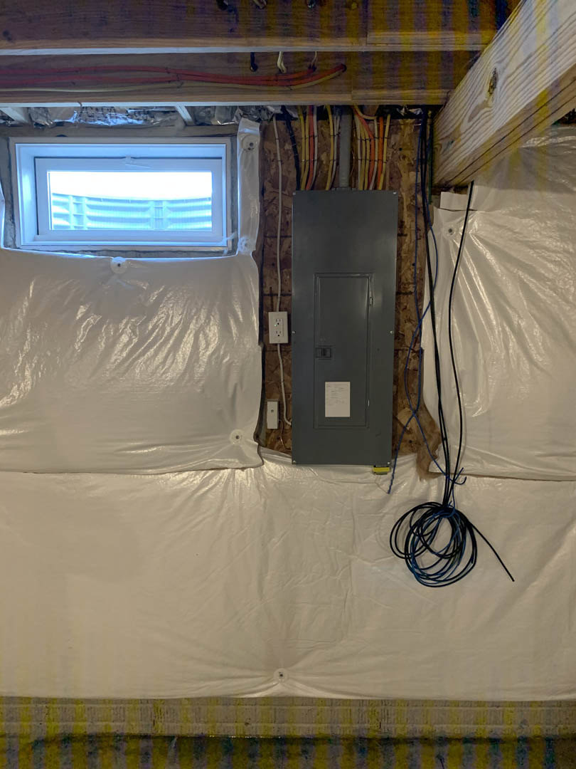 Bedroom wall with exposed wires and plastic coverings, grey metal door with lock, electrical box, plastic bag taped to window, piece of paper on black surface