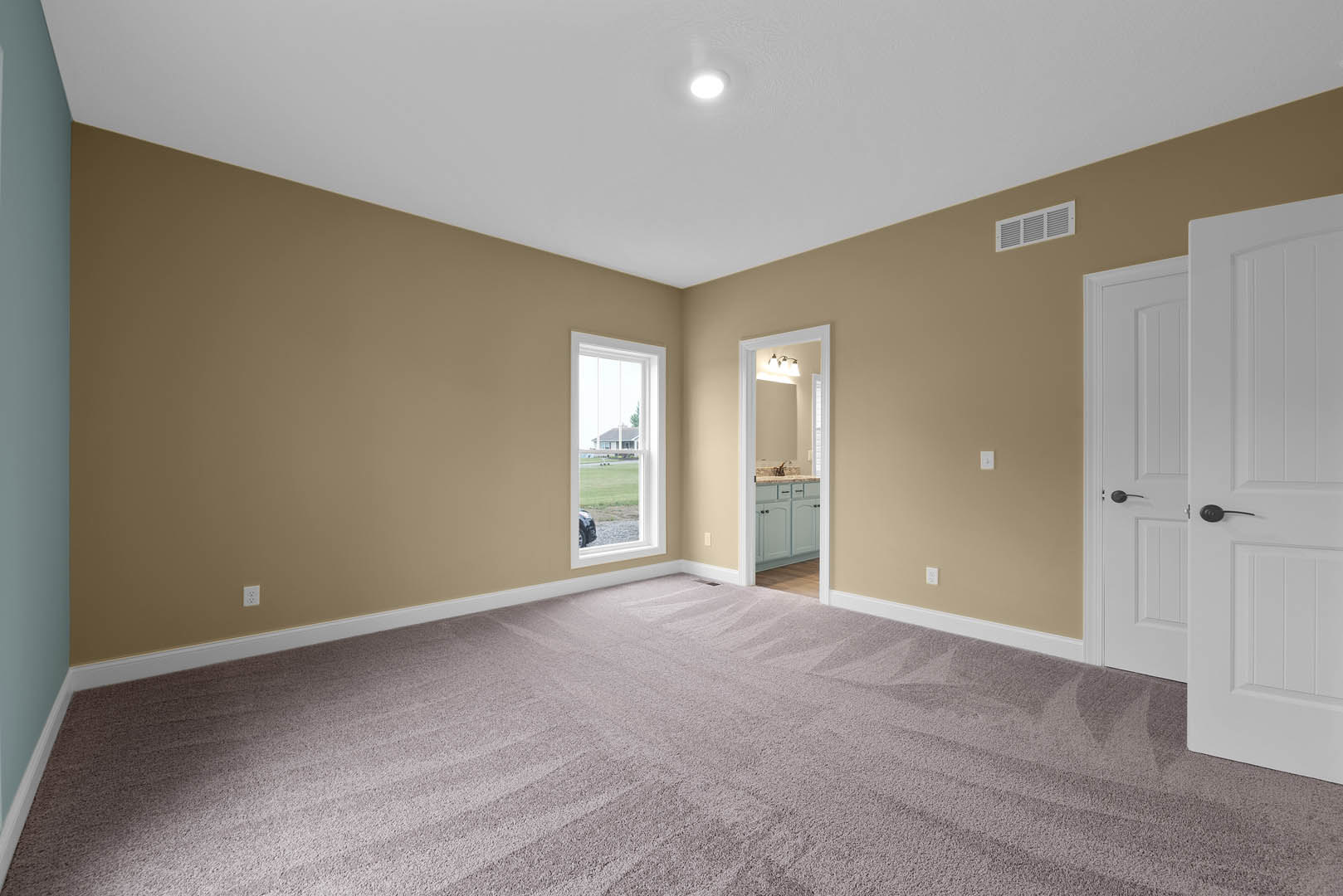 Beige-walled room with carpet flooring, ceiling light fixture, window showing neighboring house, bathroom featuring sink and mirror, close-up of brushed metal door handle