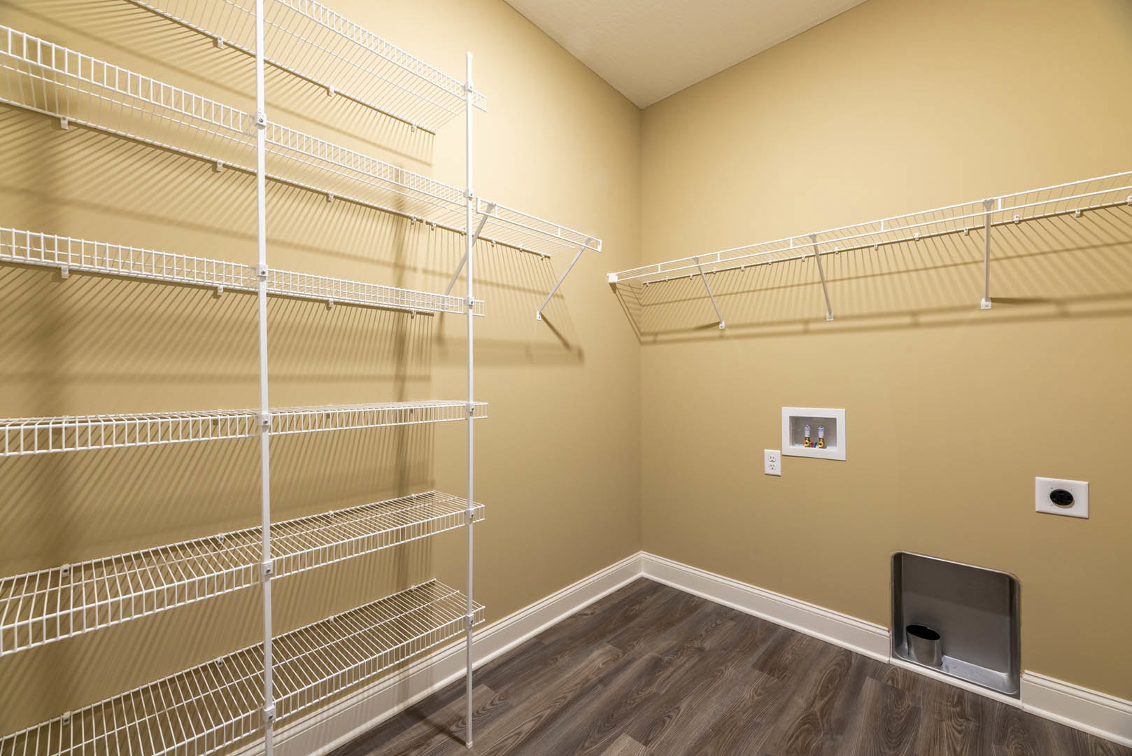 Built-in white shelves on a plaster wall, wood plank flooring, white ceiling, white shelf with hooks, exposed pipes in a recessed box, black oval outlet on wall.