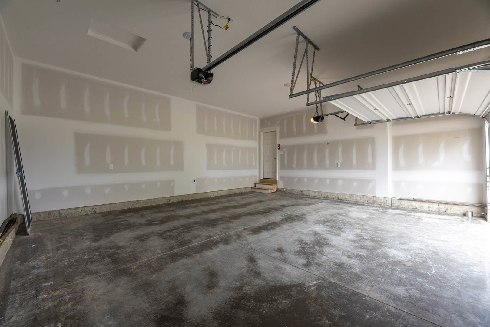 White-walled room with concrete floor, visible staircase, white door with black handle, and garage doors; close-up of stair and bag.