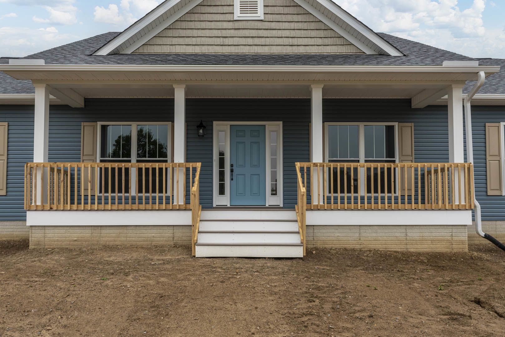 Blue front door with white trim and black handle, porch with light-colored siding, window adjacent to entry, dirt ground in foreground.