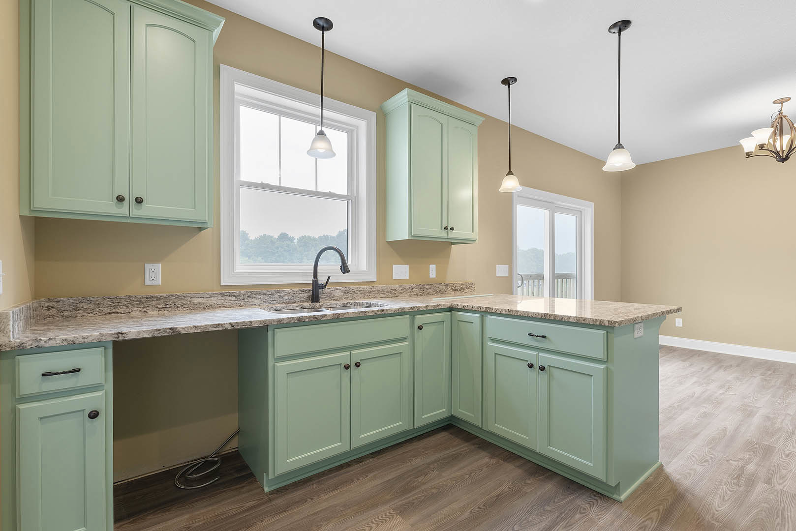 Green kitchen cabinets with marble countertops, tile backsplash, stainless steel sink and faucet, drawers and cupboards, overhead lighting fixture, metal tube visible on the floor.