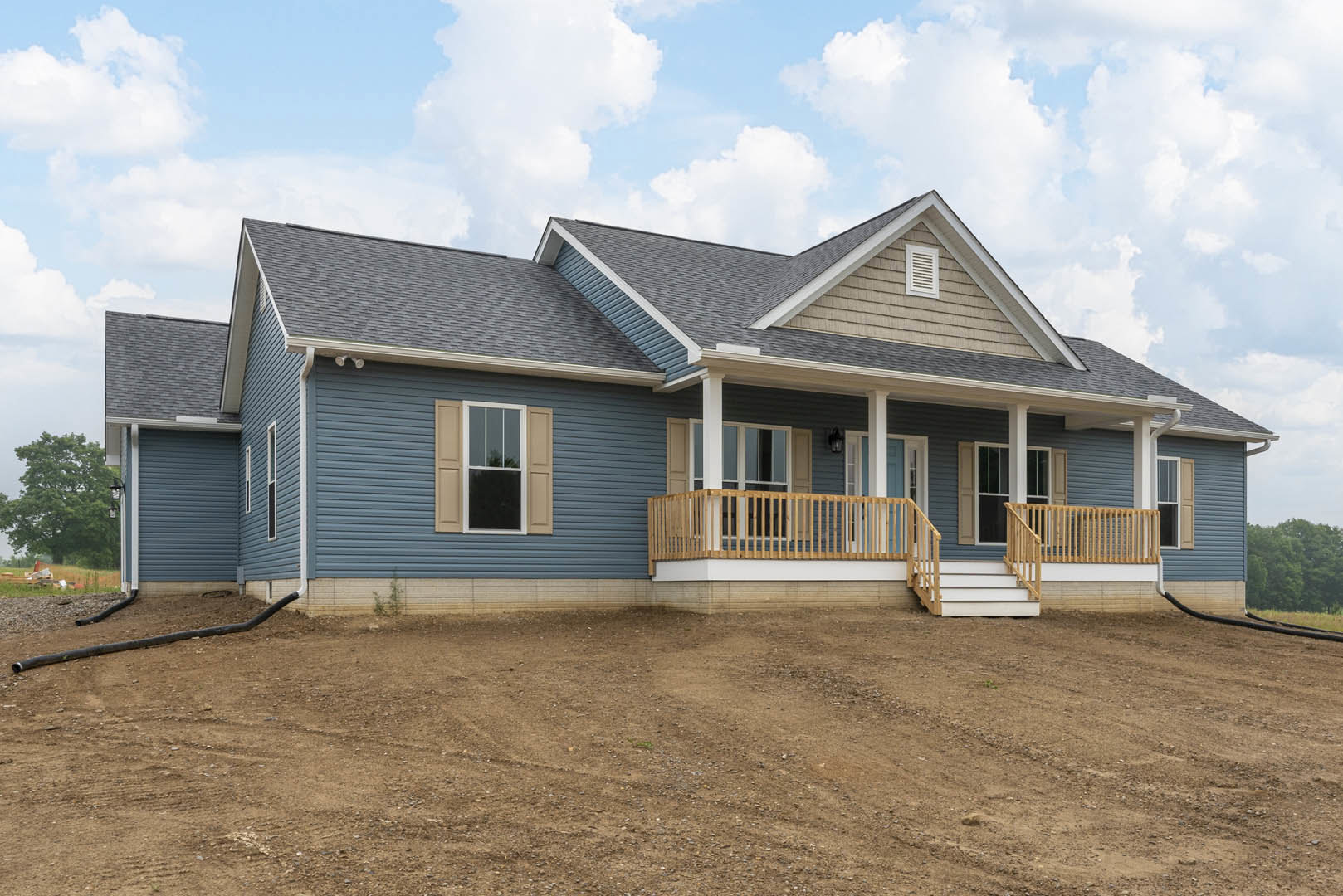 Two-story house with white-framed windows and covered porch, set beside a dirt hill and field, green-leafed tree, and black pipe on the ground