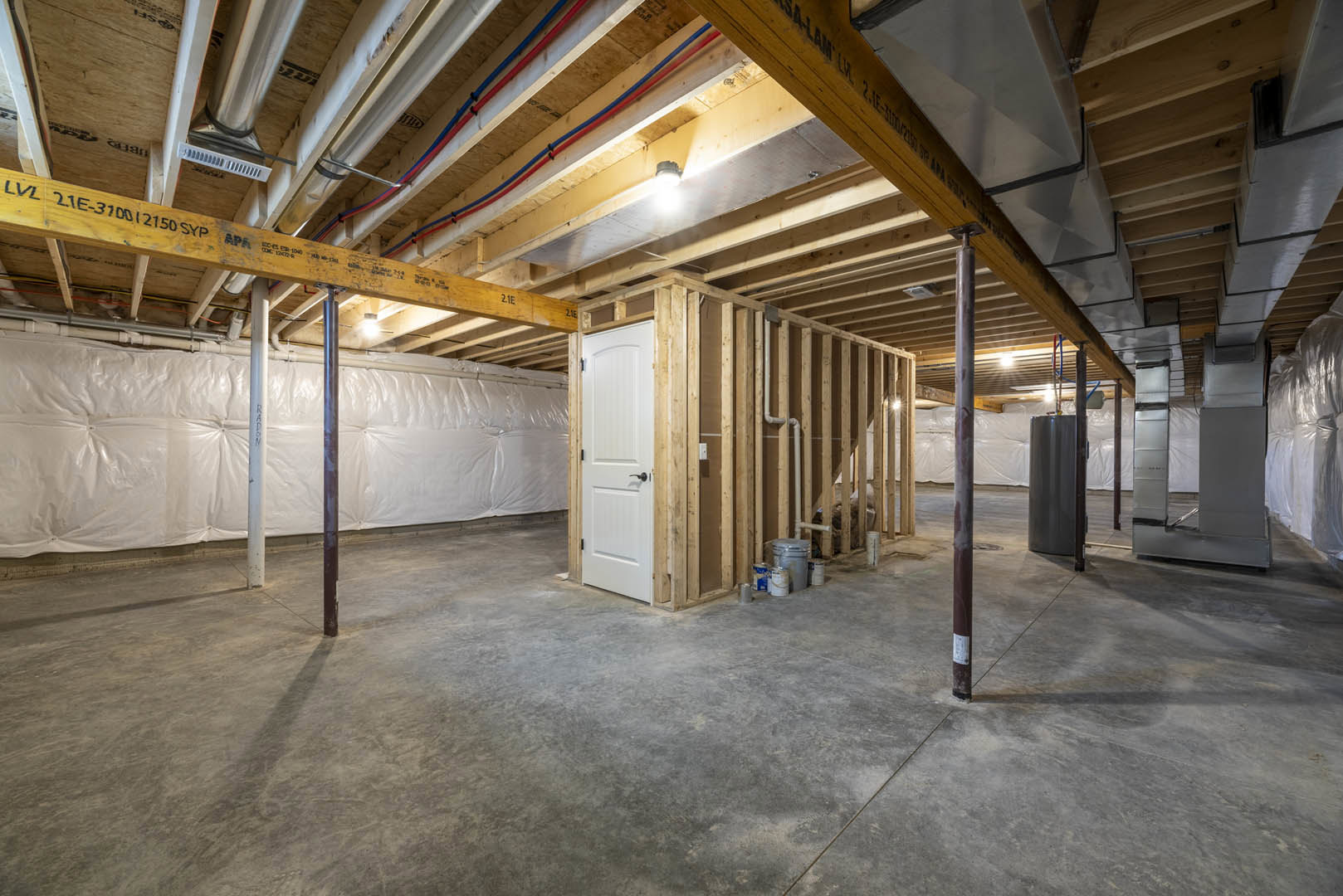 Basement room with concrete floor, white door featuring black handle, exposed yellow steel beam with black writing, white pipe along wall, metal object and paint cans near door.