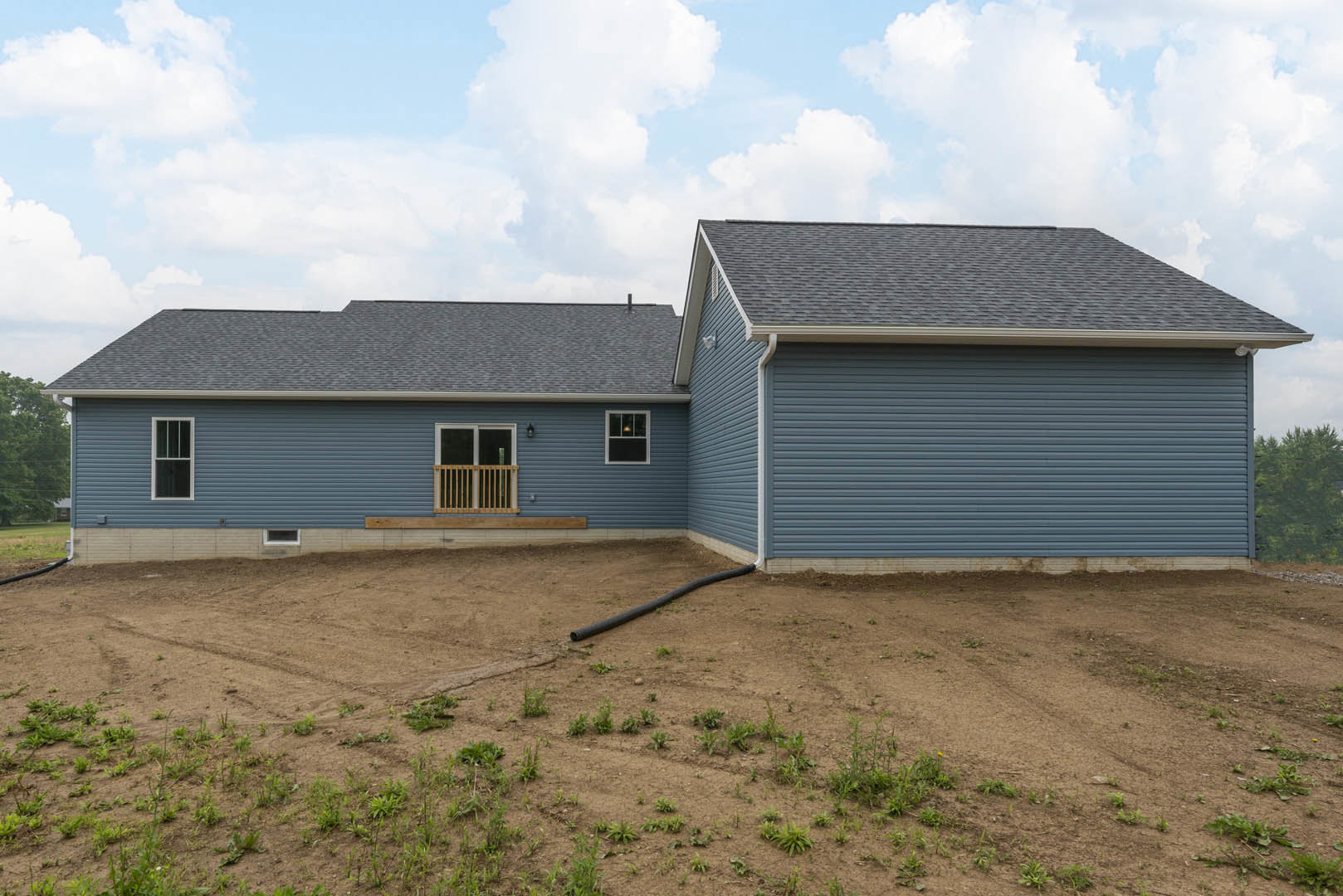 Two-story house with light siding, covered porch, upper balcony, dirt yard, black pipe on ground, illuminated window, cloudy sky