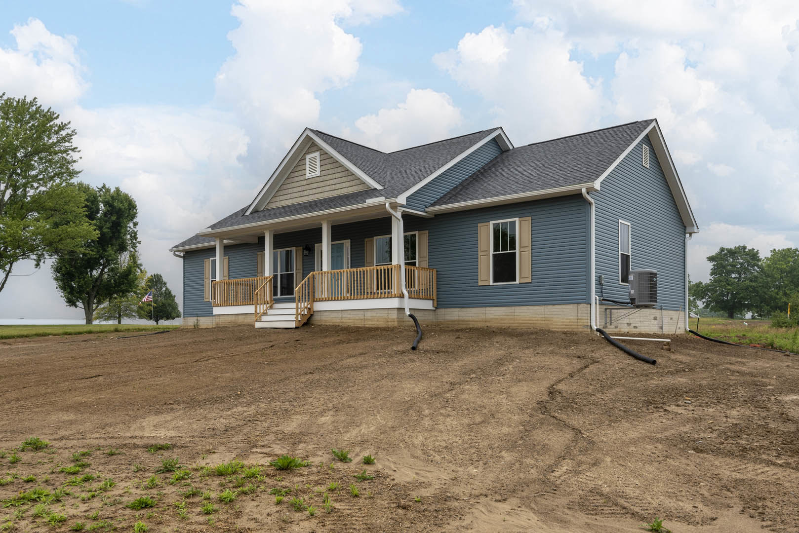 Large front porch with white-framed windows, wooden railing, and dirt hill in foreground; mature trees and cloudy sky in background