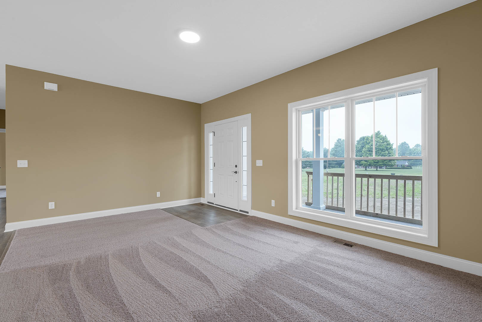Bright room with white walls, laminate flooring, a white door with glass panel, large windows overlooking a porch, ceiling light fixture, and carpeted area near the entrance.