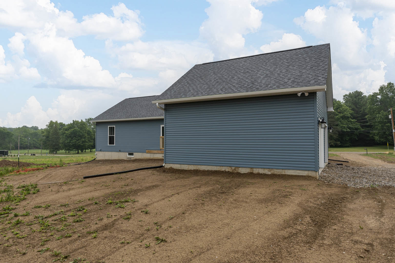 Single-story home with gray roof and white siding, surrounded by dirt field, scattered rocks, metal pipe, and mature trees under cloudy sky