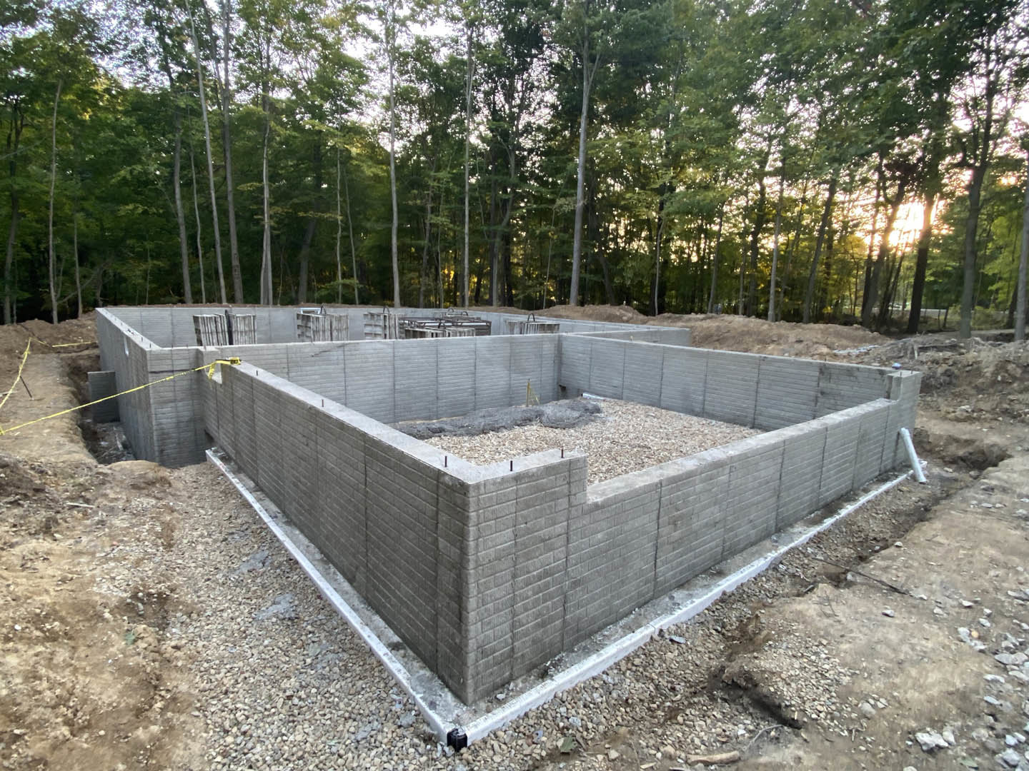 Concrete foundation surrounded by dirt and construction materials, pile of wood on the slab, trees lining the background under a clear sky