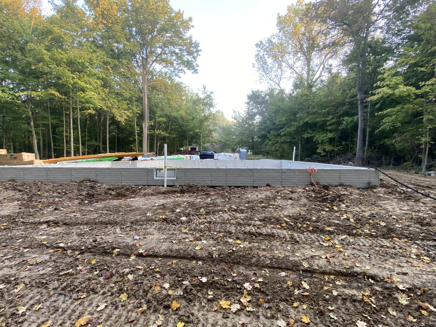 Concrete foundation slab with exposed rebar and black drainage pipe, surrounded by dirt field with scattered leaves, pile of soil, nearby asphalt road, and trees in the background