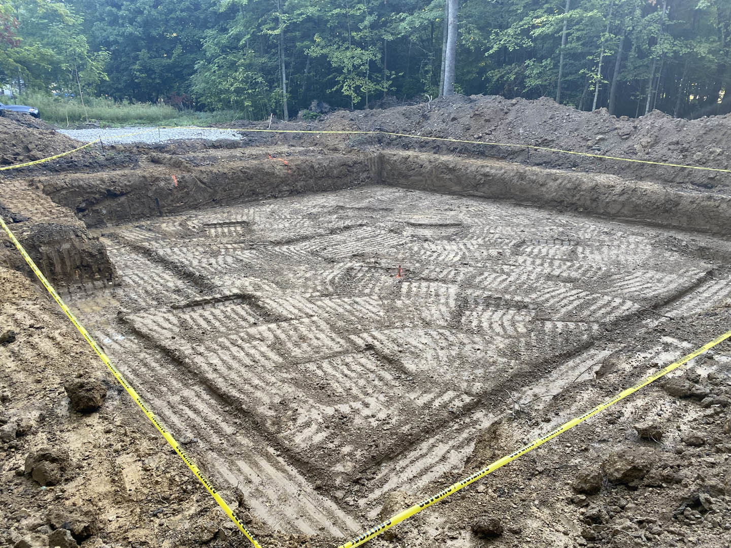 Square patch of bare soil bordered by yellow tape, surrounded by outdoor greenery and trees