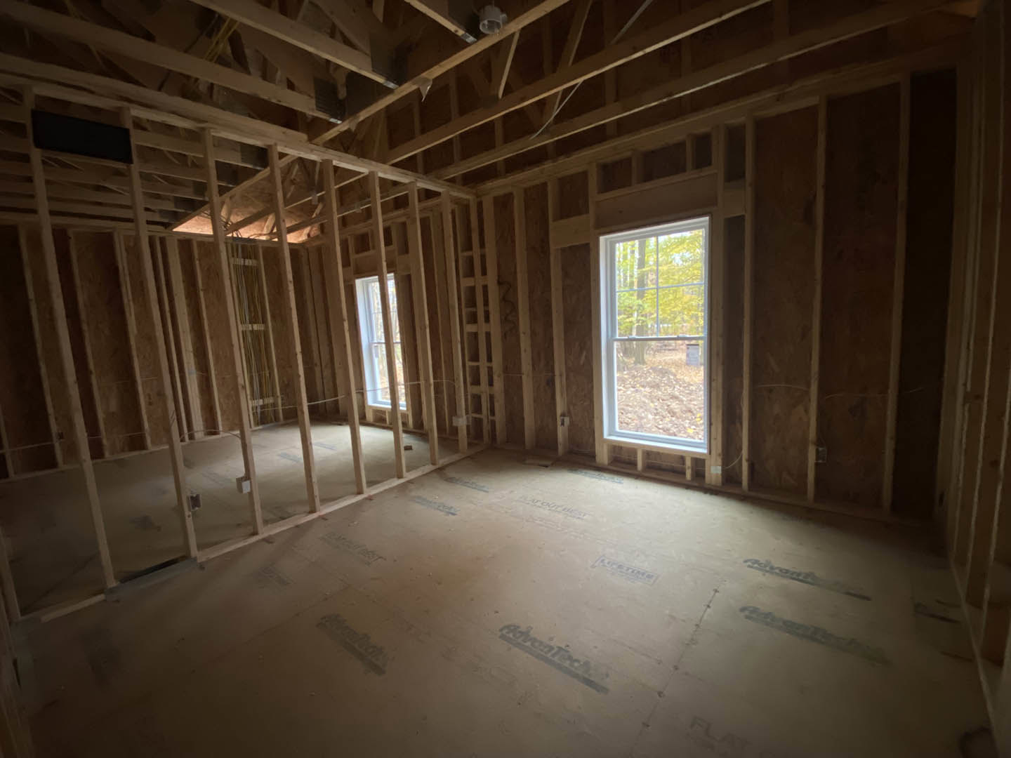 Wood-framed room under construction with exposed beams, unfinished plaster walls, large windows letting in daylight, and view of trees outside
