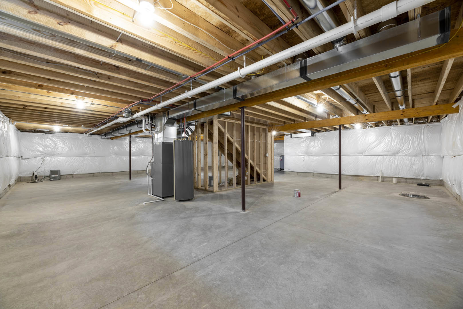 Exposed pipes along ceiling, concrete floor with wooden framing, grey rectangular object featuring white stripe, white plastic wall covering, unfinished basement interior