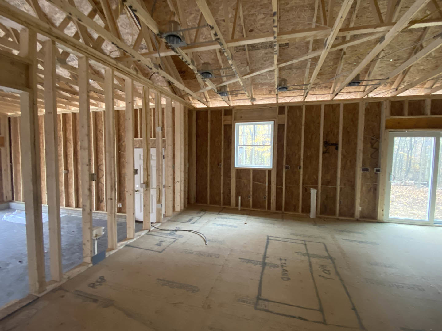 Living room with exposed wood ceiling beams, large window framed in white, glass door overlooking forest, light-colored floor featuring a black accent line.