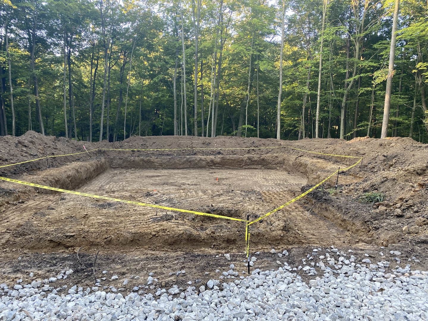 Dirt construction site bordered by yellow caution tape, scattered rocks, and surrounding trees in a wooded area
