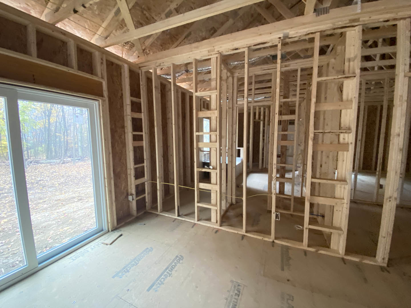 Open-concept living area with exposed wooden ceiling beams, white plaster walls, large windows overlooking trees, and light hardwood flooring