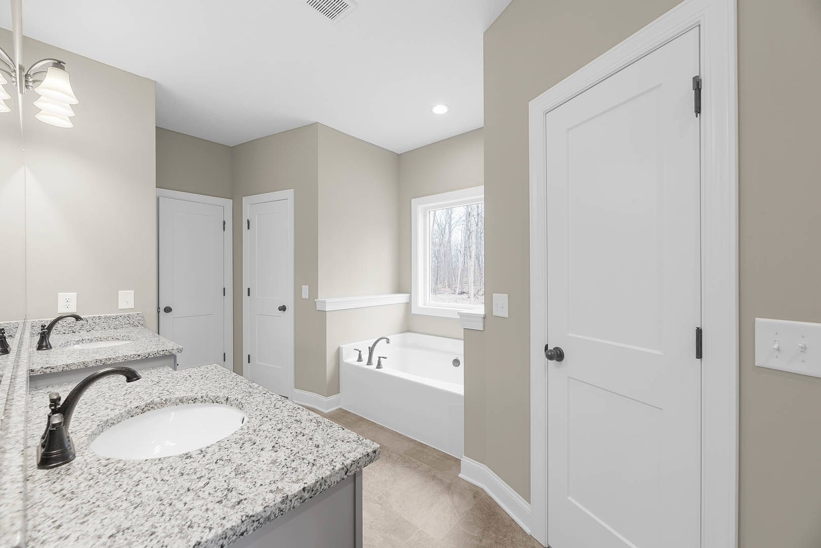 Bathroom featuring marble countertops, white sink with black speckled surface, freestanding bathtub, white door with black hardware, large window overlooking trees, and light tile