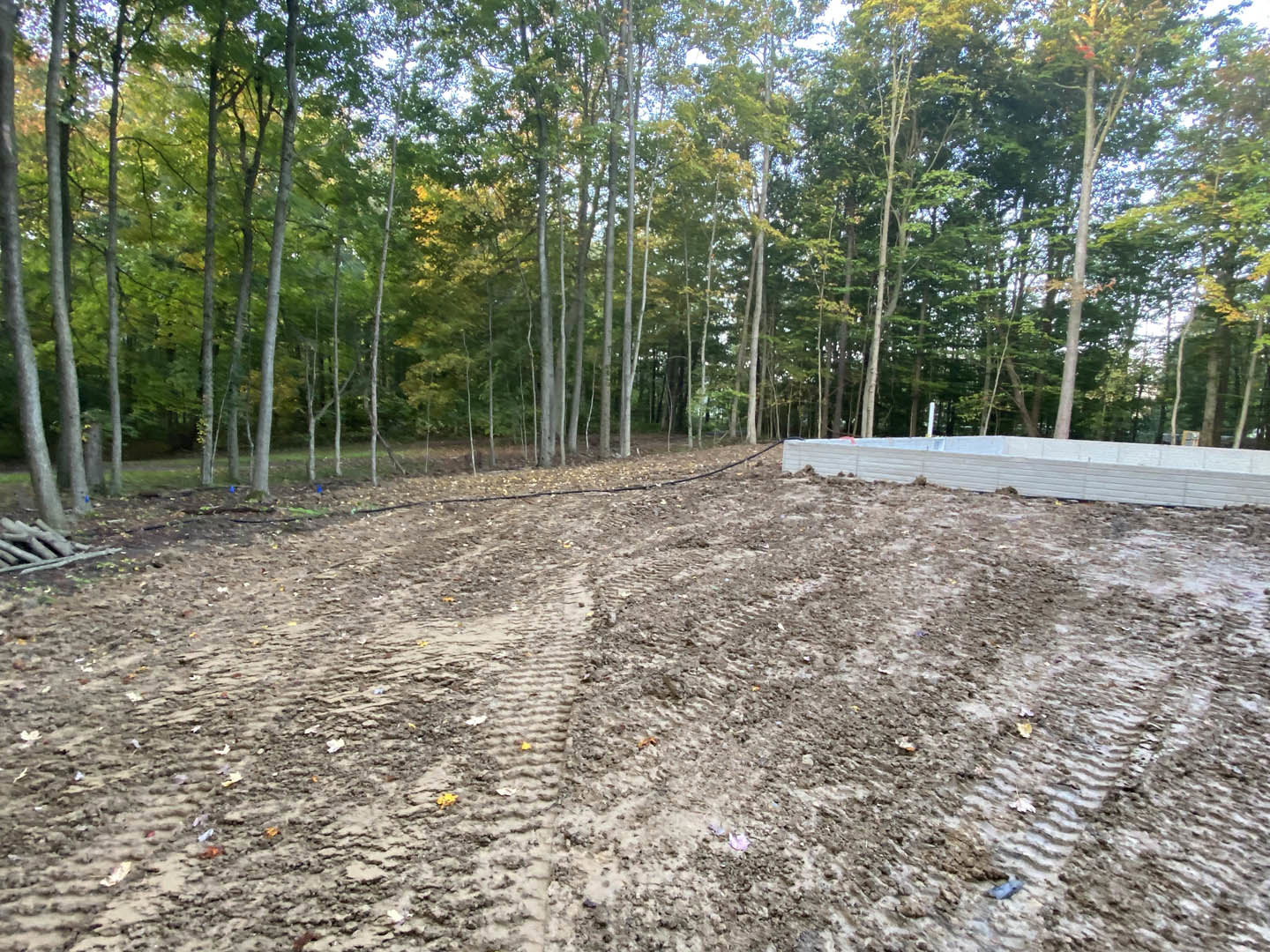 Dirt field with tire tracks, pile of logs, white rectangular object with cover, white wall, and tall deciduous trees in the background