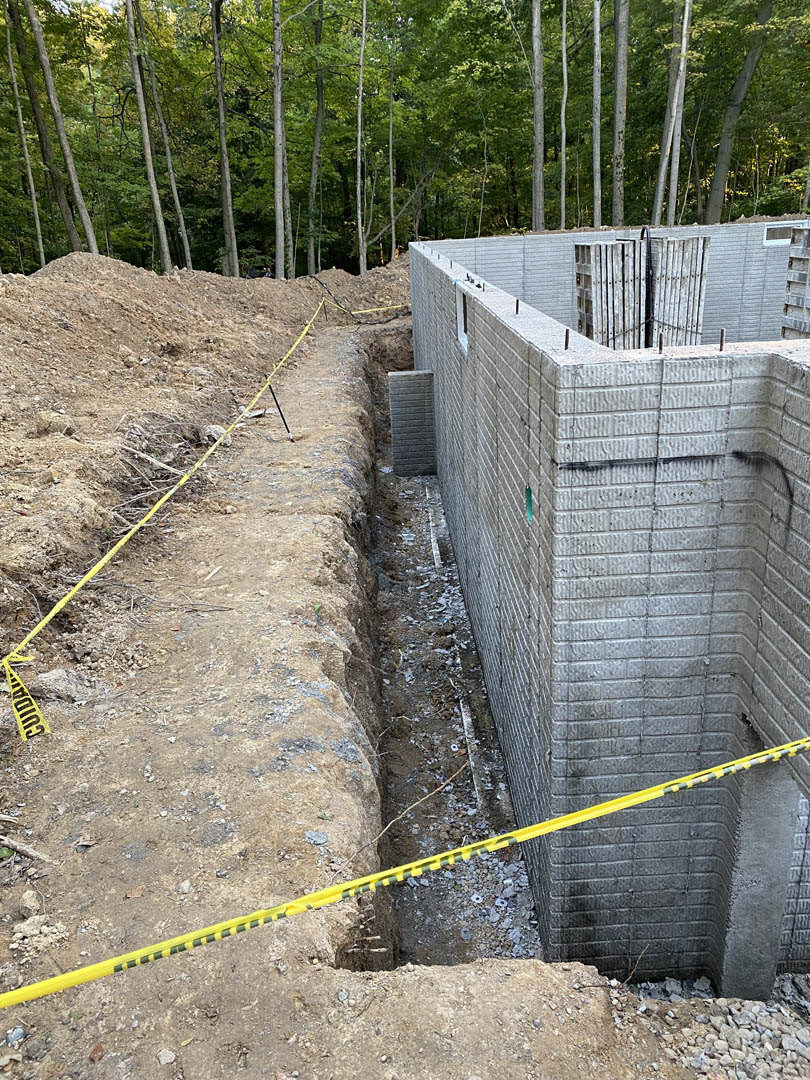 Concrete foundation wall bordered by yellow caution tape on dirt ground, surrounded by trees in wooded construction site