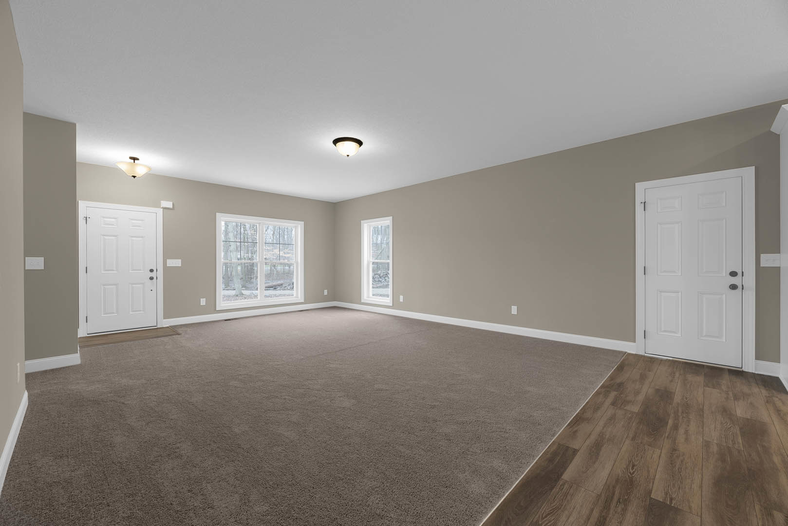 White paneled door with black handle, multiple windows overlooking trees, wood flooring partially covered by a light carpet, smooth white walls and ceiling.
