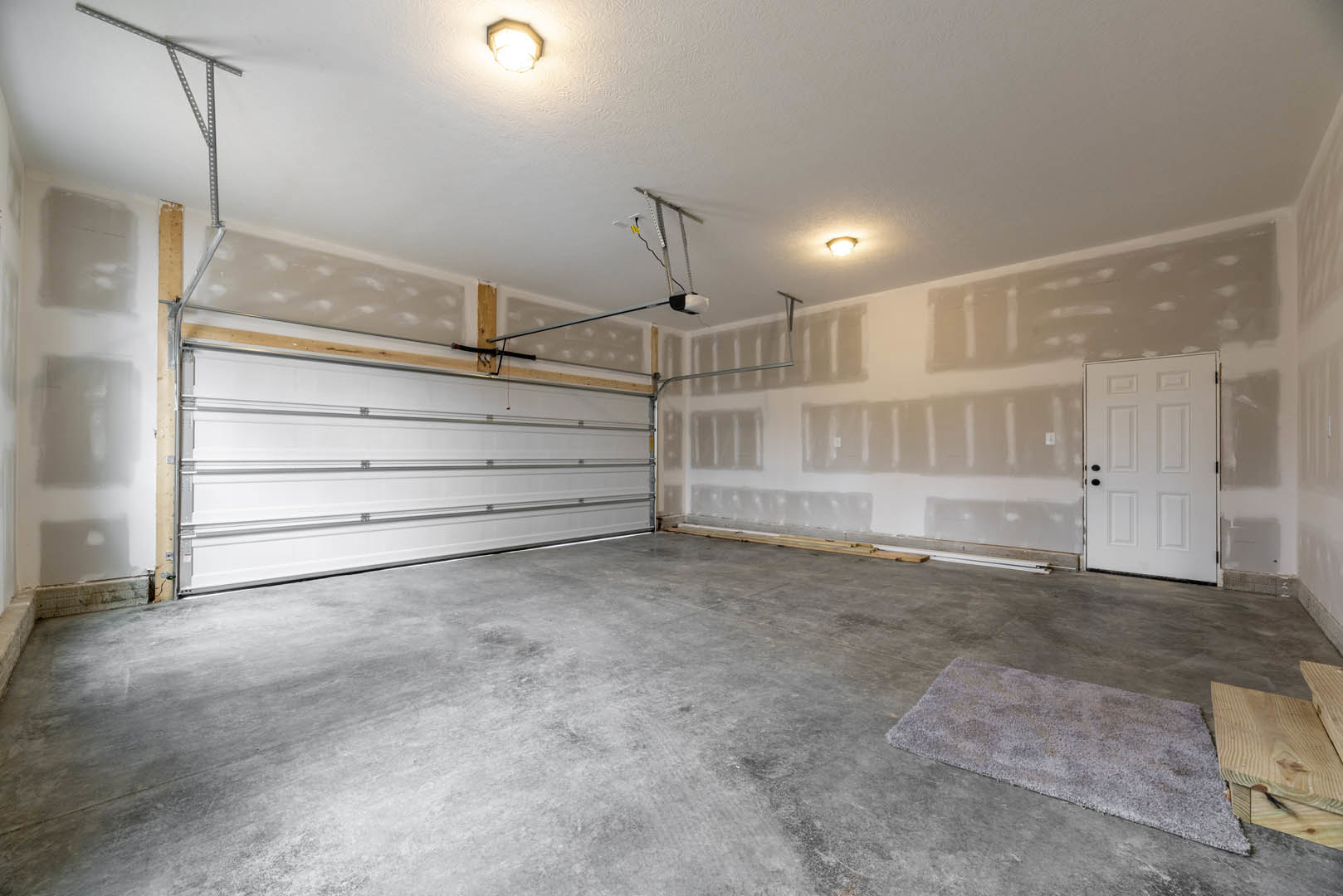 Garage interior with white paneled door featuring black handles, concrete floor, neutral plaster walls, ceiling light fixture, and a patterned rug near the entrance.