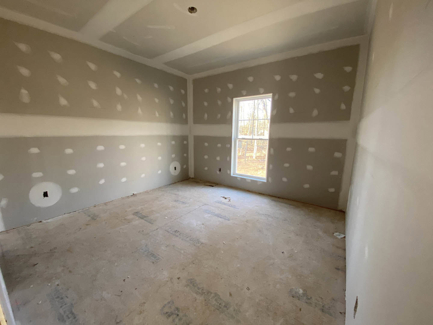 White plaster walls, large window overlooking forest, light wood flooring, black object resting on white windowsill, wall with small holes visible near floor
