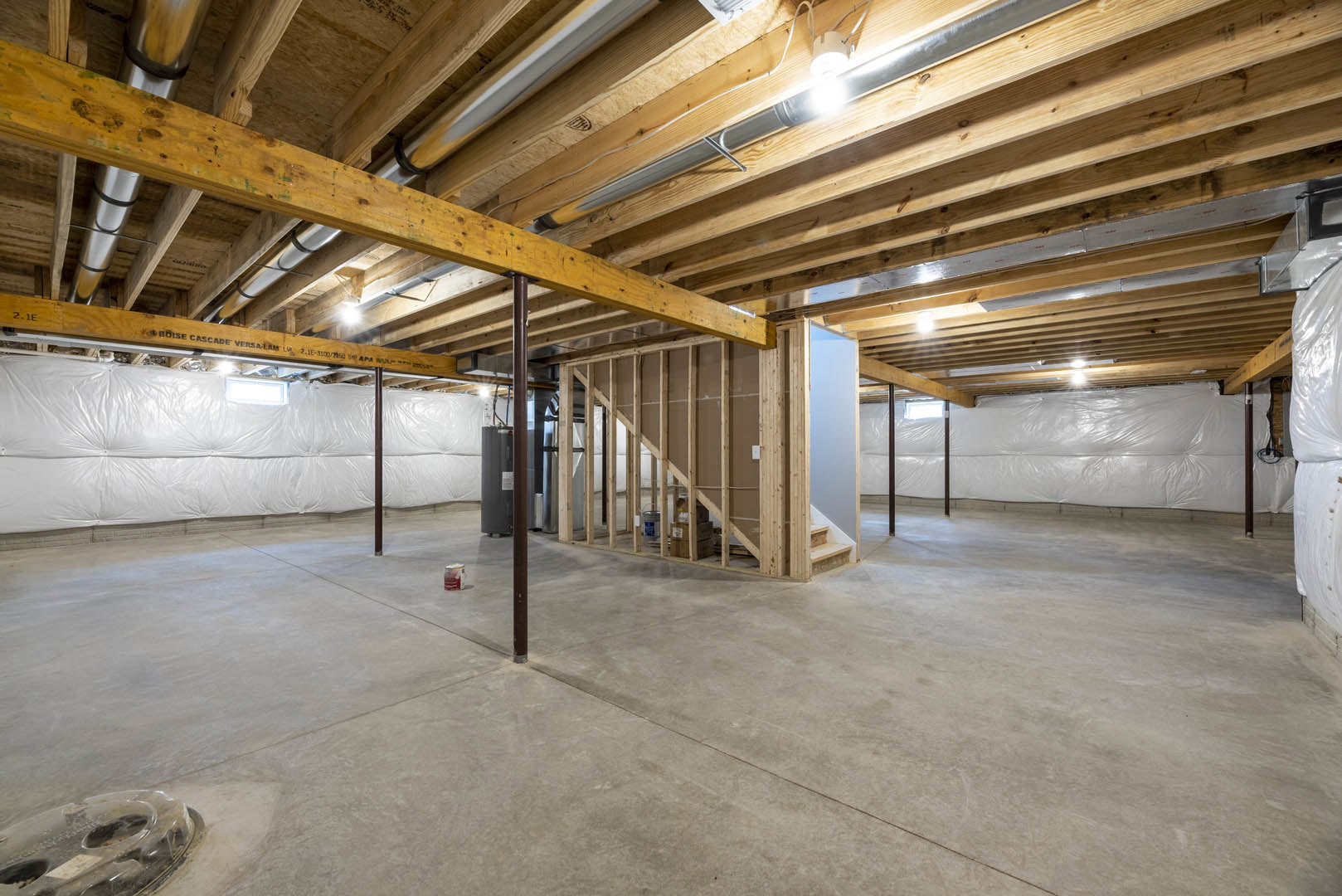 Basement room with exposed wood ceiling beams, concrete floor, steel pipe along ceiling, white plastic wall covering, and staircase against one wall.