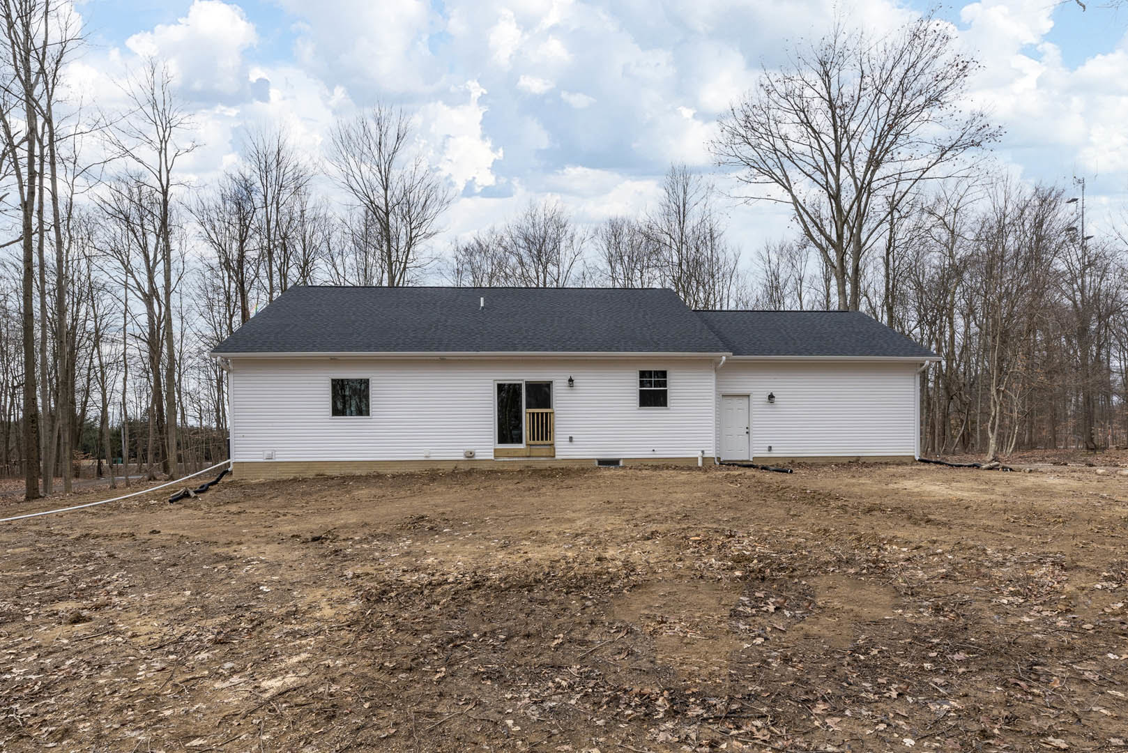White house with black roof, white-framed windows, wooden railing, dirt field with scattered leaves, trees and cloudy sky in background