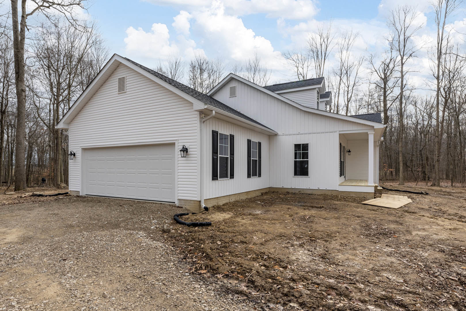 White house with multi-pane windows, attached garage, concrete driveway, exposed dirt area with black pipe, white siding, overcast sky, trees in background