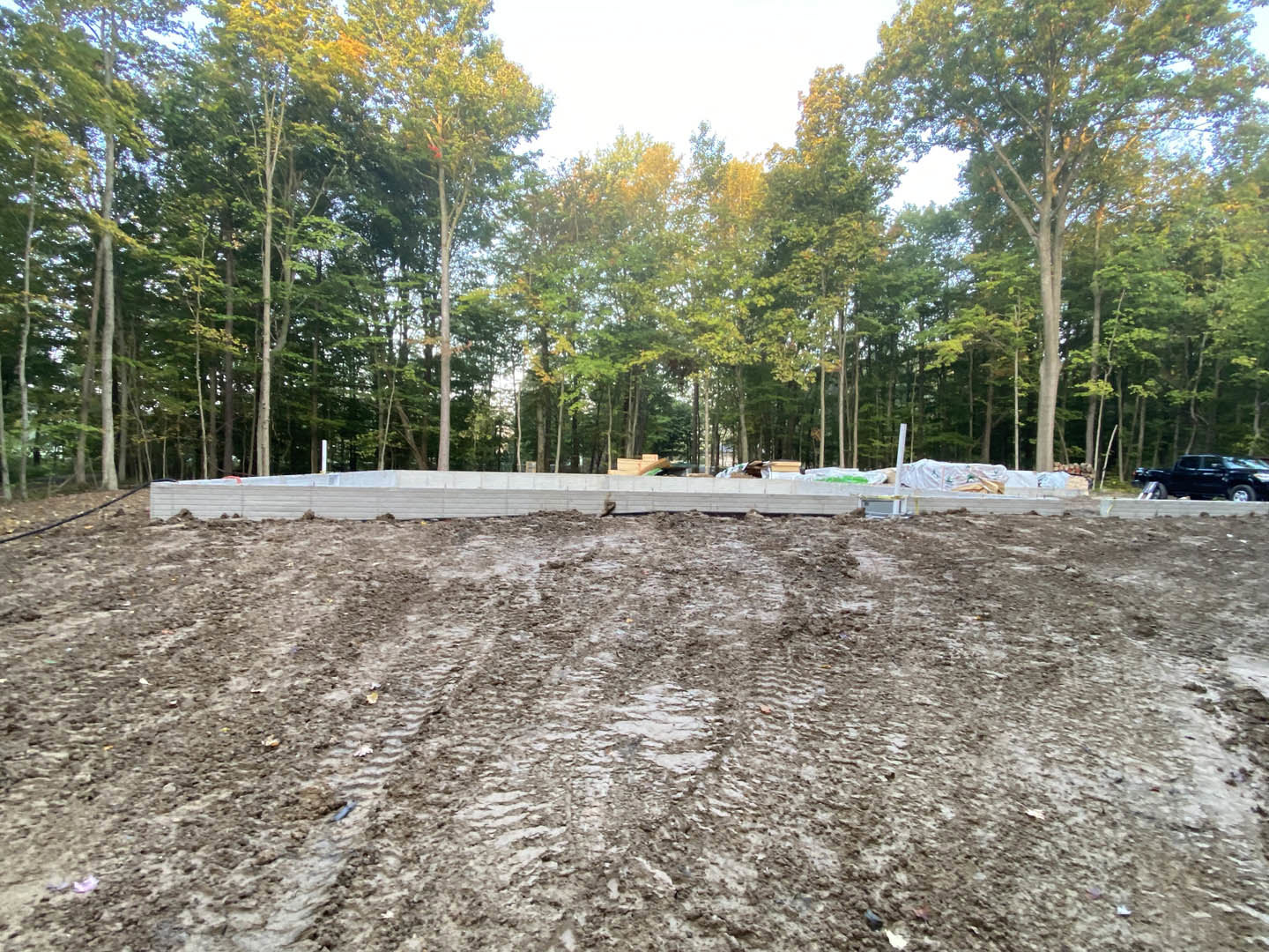 Dirt field bordered by trees, muddy ground with scattered wood, partial view of a black truck, white wall and fence in background